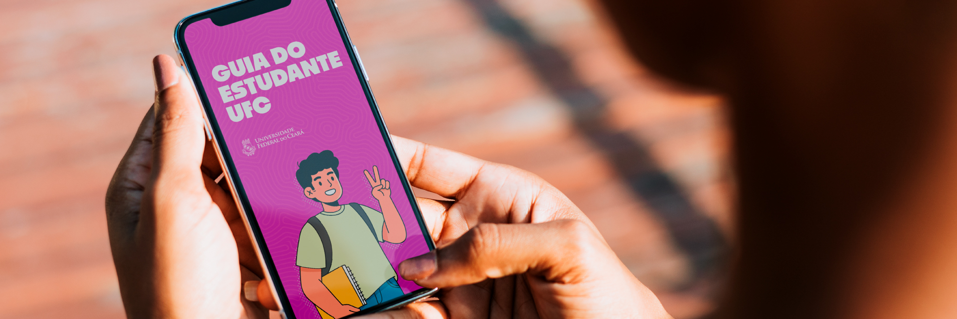 Mockup de pessoa segurando um celular com a imagem da capa do Guia do Estudante UFC na tela; a imagem do Guia possui fundo lilás e o desenho de um aluno segurando um caderno e mochila